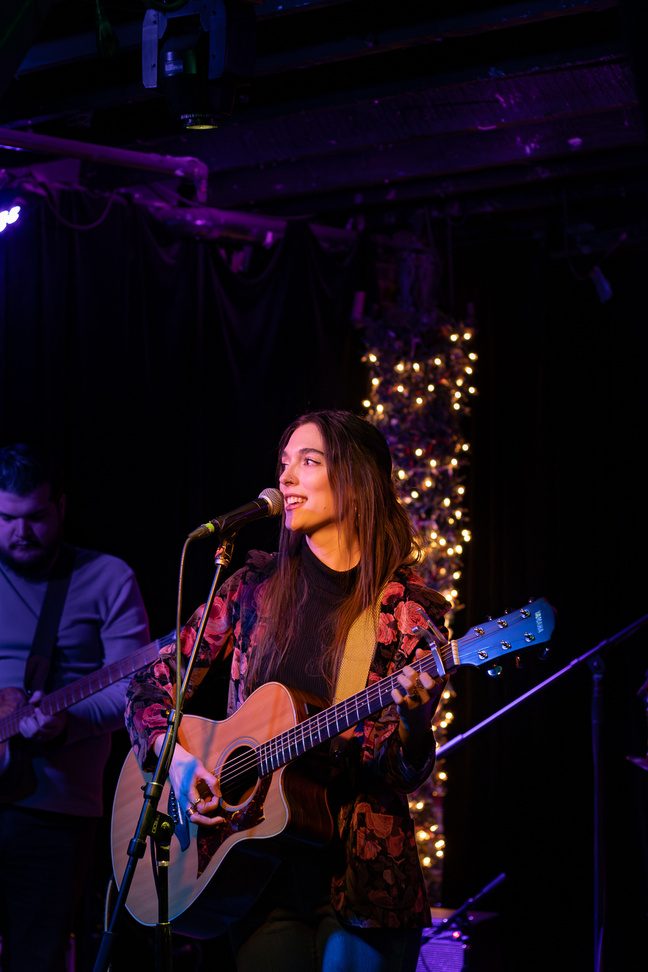 Caroline Adele singing at a microphone with an acoustic guitar, string lights behind her