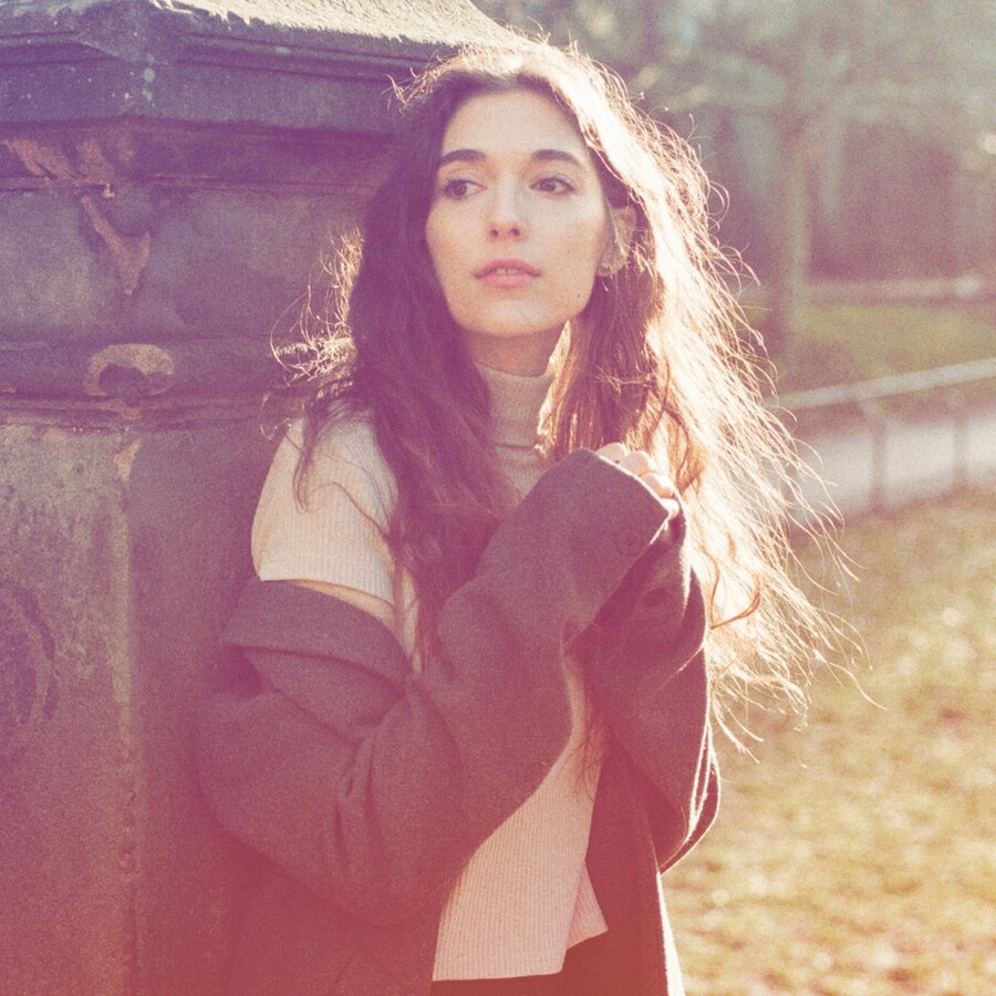 Caroline in a dark coat, sun behind her hair, beside a stone pillar